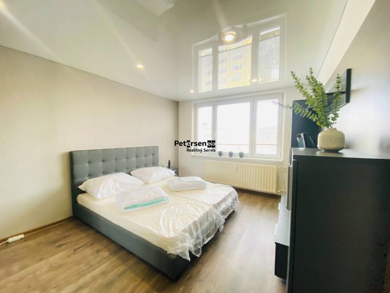 A bedroom in a three-room apartment with a wood-patterned floor and a large window.