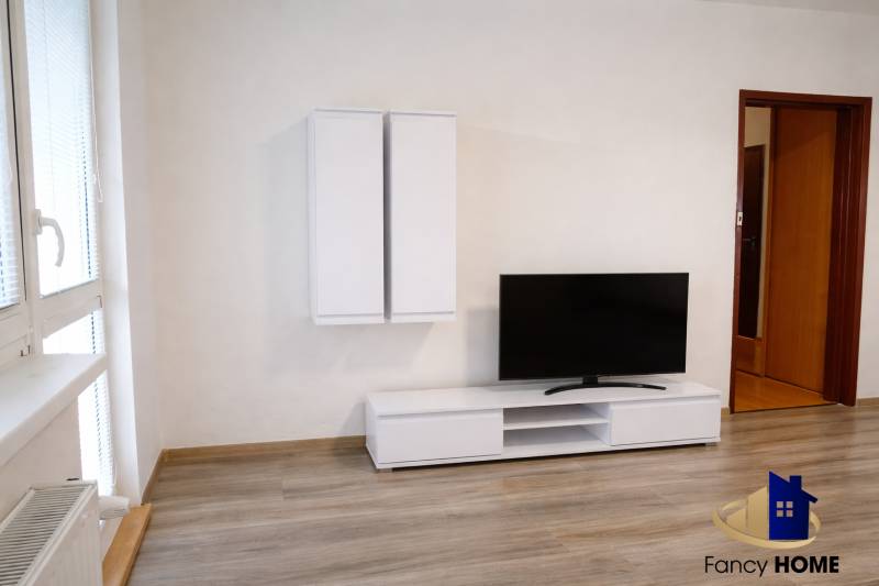 Living room in a three-room apartment with wood-patterned flooring, a television, and white furniture.