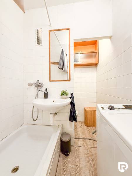 Bathroom with a shower, sink, washing machine, and floor with a wooden decor in a studio apartment.
