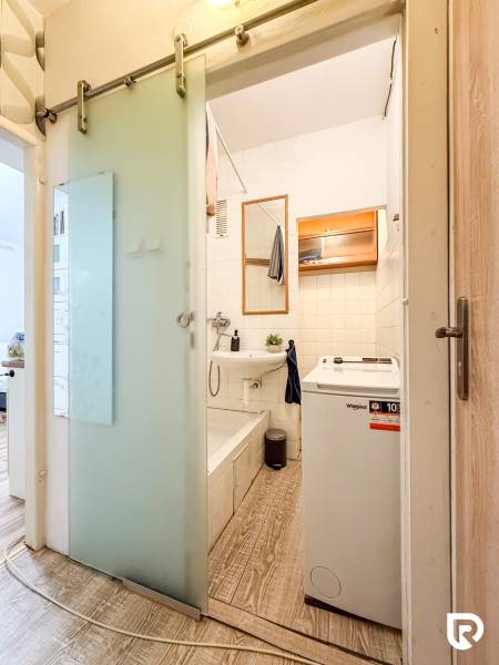 A bathroom in a studio apartment with a sink, bathtub, washing machine, and a floor with a wooden decor.