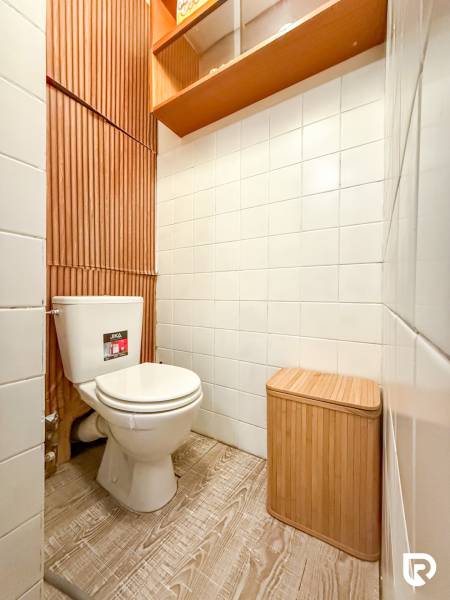 A bathroom in a studio apartment with a toilet, white tiles, wooden decor, and storage shelves.
