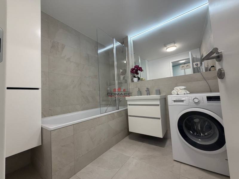 Bathroom in a 2-room apartment with a bathtub and a washing machine, light tiles and a sink.