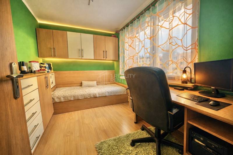 A room in a 4-room apartment with a desk, a bed, and a floor with a wood decor.