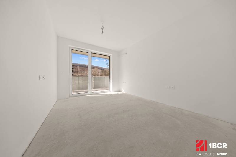 A spacious room with a gray floor and a large window in a 4-room apartment.
