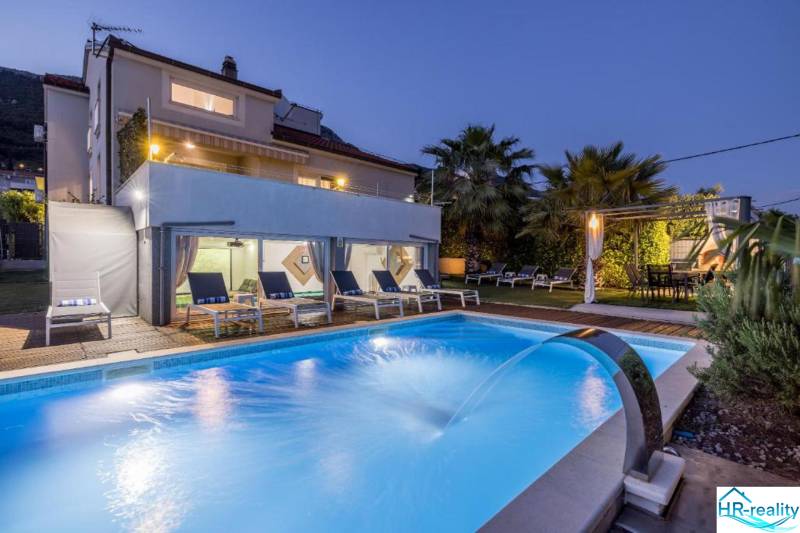 Villa in Kaštela with a lit pool, loungers, and palm trees in the garden at dusk.