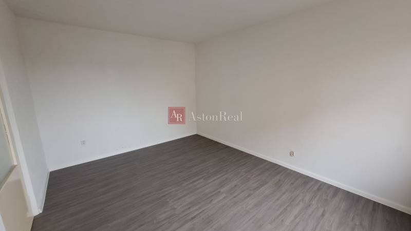 An empty room in a one-room apartment with white walls and a wooden-patterned floor.