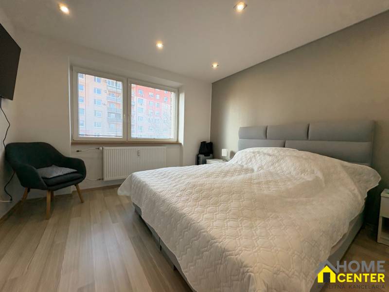 A bedroom in a 2-room apartment with a wood-patterned floor, a comfortable armchair, and a large window.