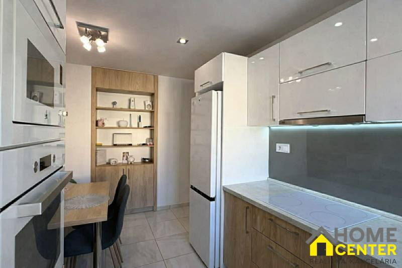 A kitchen in a 2-room apartment with white cabinets, a table, and wooden decor shelves.