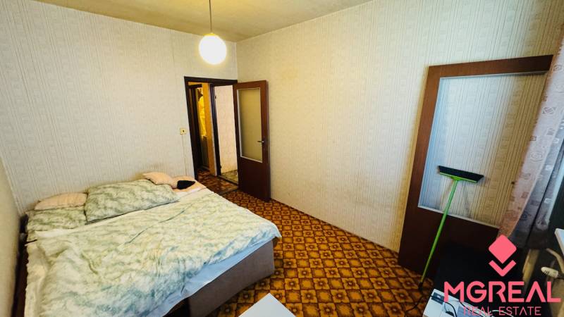 A bedroom with a carpet, a bed, and light-colored walls in a 2-room apartment.