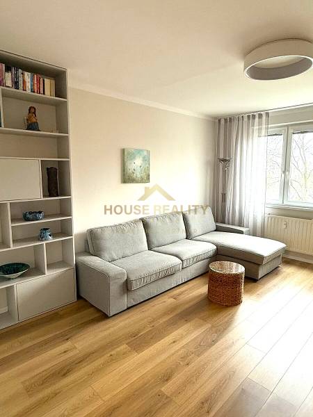 Living room of a three-room apartment with a gray sofa, a shelf, and a wooden-patterned floor.