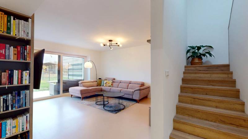 Interior of a family house: sofa, bookshelf, stairs with wood-patterned flooring, and a plant.