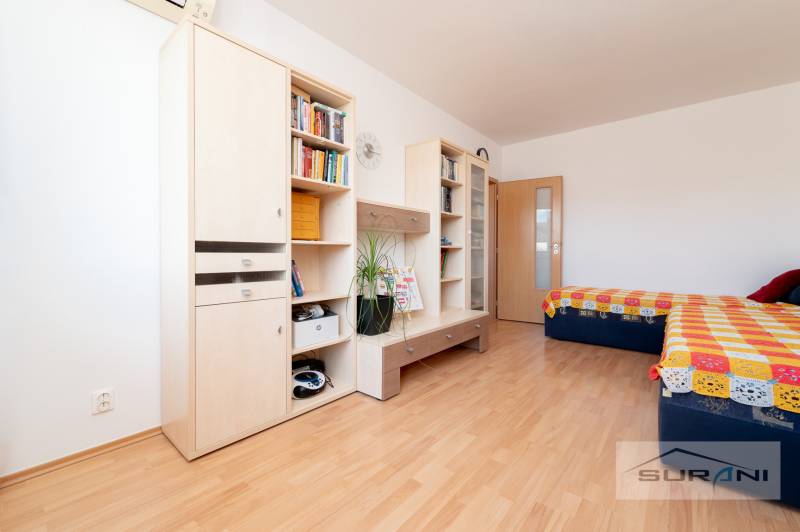 A room with a wooden decor floor contains furniture, shelves, a double bed, and a colorful blanket.