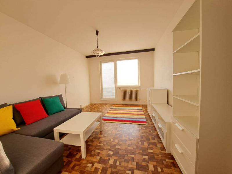 Living room in a two-room apartment with a sofa, white furniture, colorful pillows, and a wooden-patterned floor.