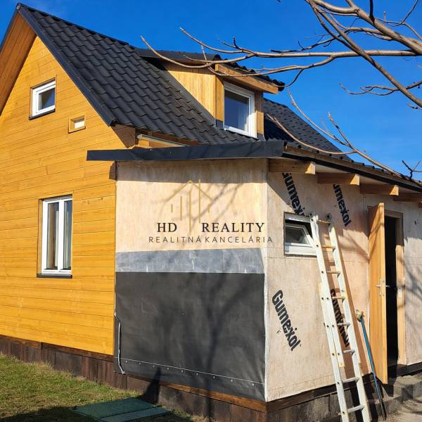 A cottage in Kráľovce Krnišov with wooden cladding and an unfinished extension against the backdrop of a blue sky.