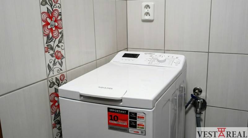 A washing machine in a studio apartment, floral patterned tiles, and a white socket on the tiles.