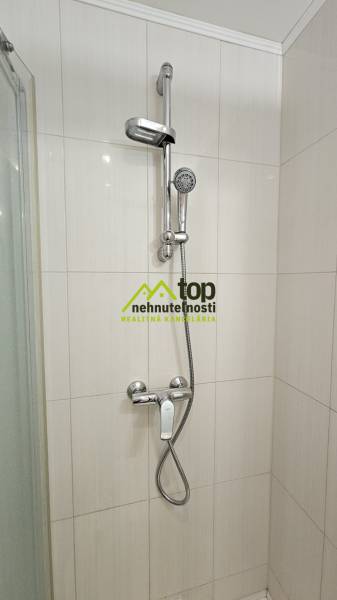 A shower corner in a studio apartment with white tiles and a chrome shower faucet.