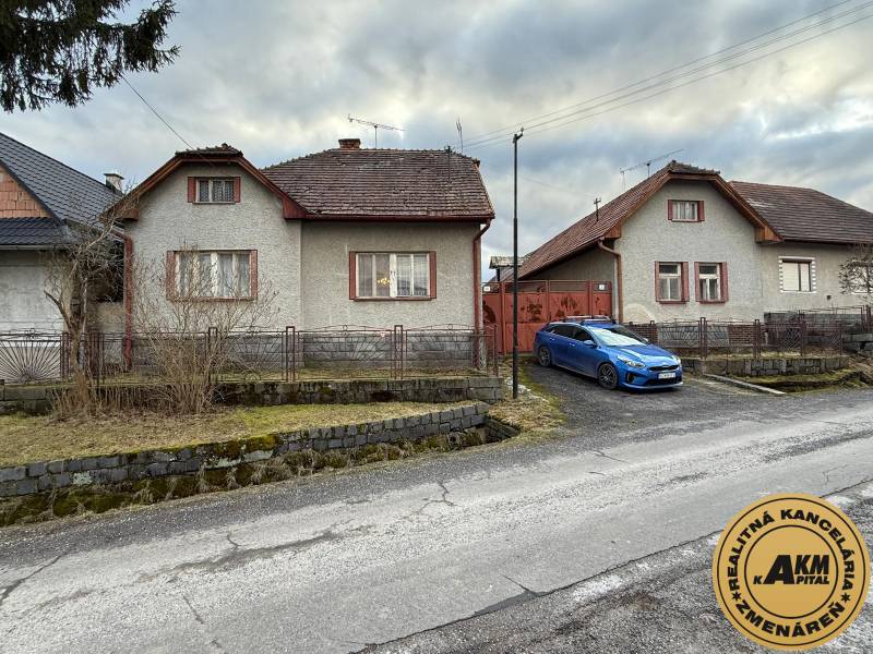 A family house in Babiná with two neighboring houses, a parked car, and a stone fence.