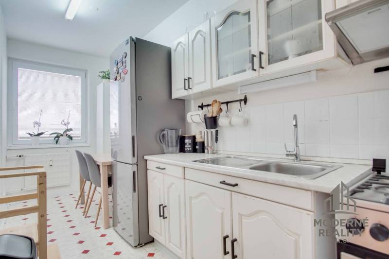 A kitchen in a 3-room apartment with white cabinets, a dining table, and a large refrigerator.