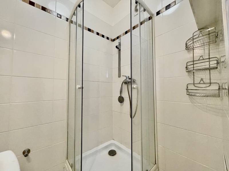 A shower corner in the bathroom of a one-room apartment with white tiles and glass doors.