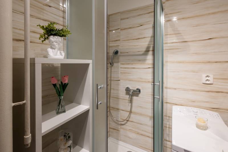 Bathroom with an enclosed shower, a shelf with decorations, and a washing machine in a 2-room apartment.