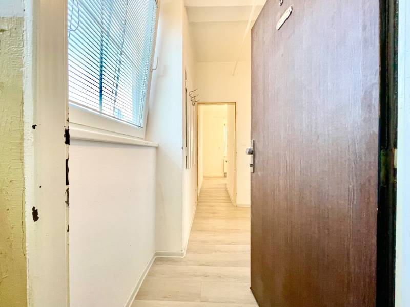 A hallway with a wooden decor floor, white walls, a large window with blinds.
