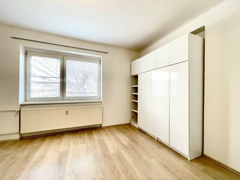 A room with a large window, a wardrobe, and a floor with a wooden decor in a studio apartment.