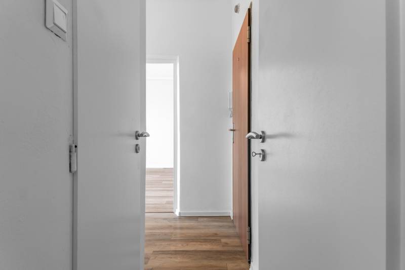 Biely interiér 1-izbového bytu s podlahou s dreveným dekorom a otvorenými dverami.