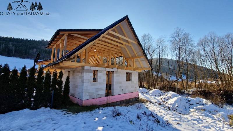 An unfinished cabin on the street in Mýto pod Ďumbierom, surrounded by a snowy landscape and trees.