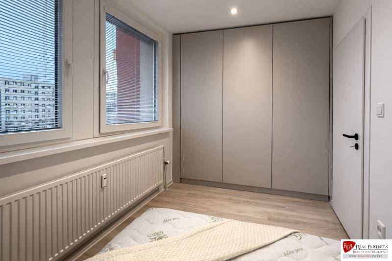 Bedroom of a two-room apartment with a built-in wardrobe, large window, and wooden decor flooring.