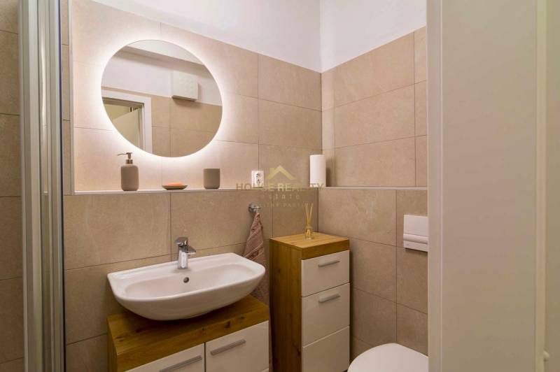 Compact bathroom in a studio apartment with a sink, illuminated mirror, and light-colored tiles.