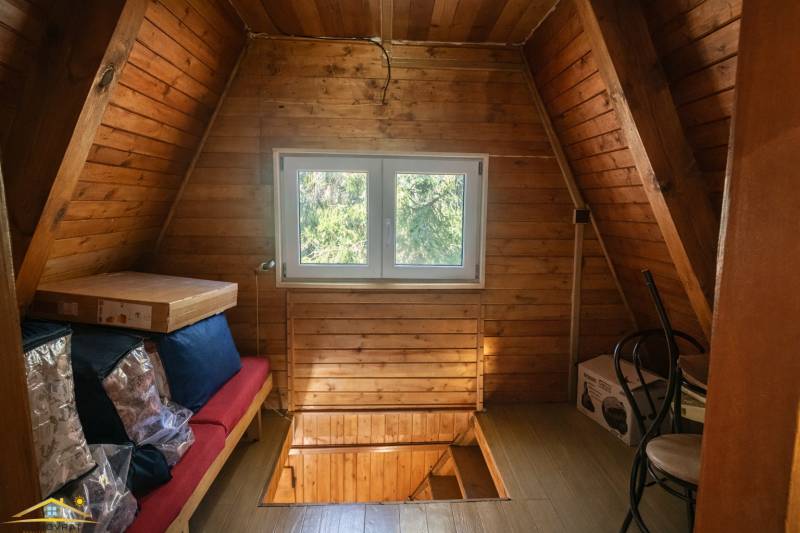 The attic of the cottage with a window, a floor with a wooden decor, boxes, and a chair.