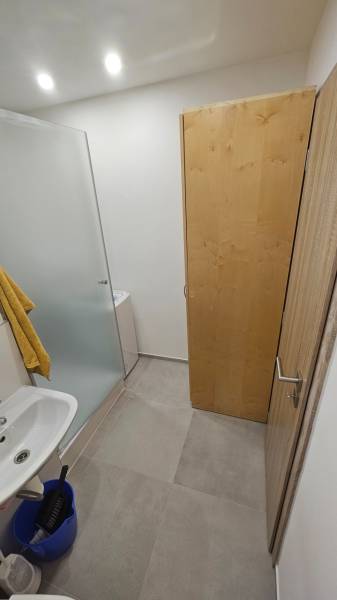 A bathroom in a studio apartment with a sink, shower, and cabinet on light-colored tiles.