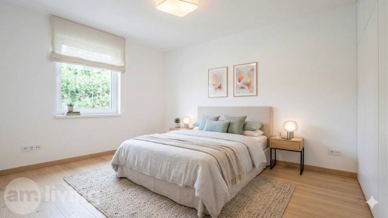 A bright bedroom with a wood-patterned floor, a bed with a light bedspread, and paintings on the wall.