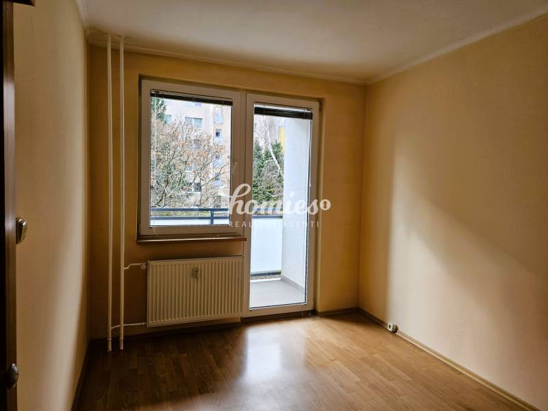 An empty room in a 3-room apartment with a wood-patterned floor and glass doors leading to the balcony.