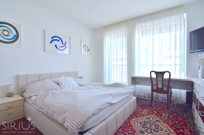 A bedroom in a 3-room apartment with a bed, a table, and artworks on the wall.