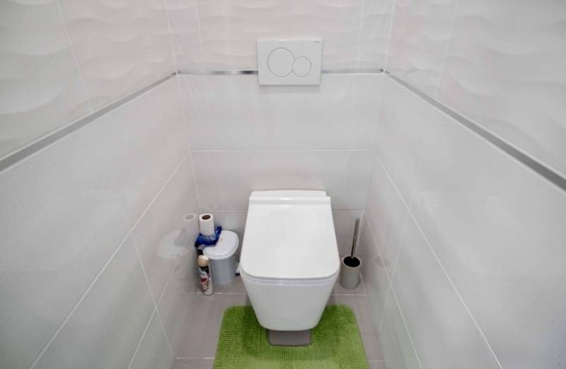A toilet with white tiles and a green bathroom rug in a 3-room apartment.