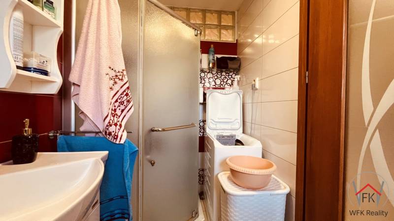 A bathroom in a 3-room apartment with a sink, shower, washing machine, and shelf.