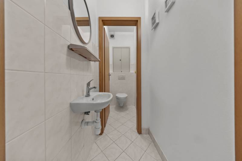 A narrow corridor with an entrance to the toilet, white tiles, a sink, and a mirror in a 4-room apartment.