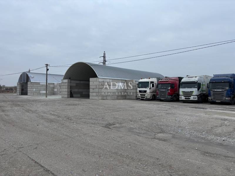 Warehouses and halls in Ladce with parked trucks on a gravel parking lot.