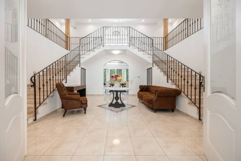 A spacious hall of a family house with a dominant staircase, brown furniture, and flowers.
