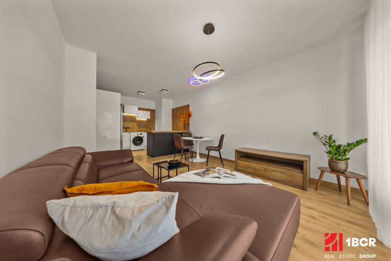 Living room in a 2-room apartment with a leather sofa, wooden decor flooring, and a dining table.