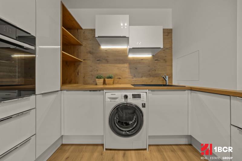 A kitchen with white cabinets, a sink, a washing machine, and wooden decor in a 2-room apartment.
