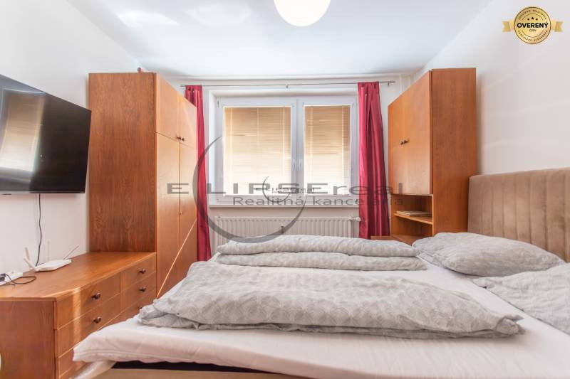 A bedroom in a studio apartment with a bed and wooden furniture, a TV on the wall.