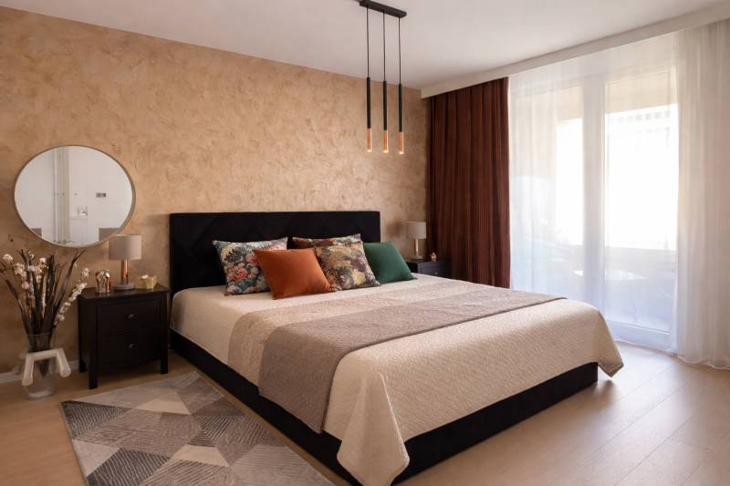 Bedroom in a two-room apartment with a large bed, mirror, and wooden decor flooring.