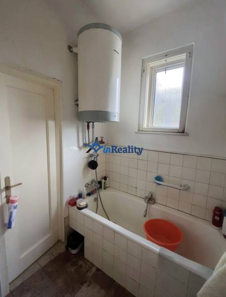 A bathroom in a family house with a bathtub, electric boiler, window, and floor with wooden decor.