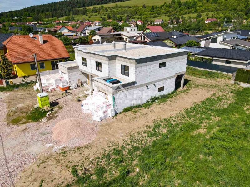 A family house under construction in Harichovce surrounded by other houses and green hills.