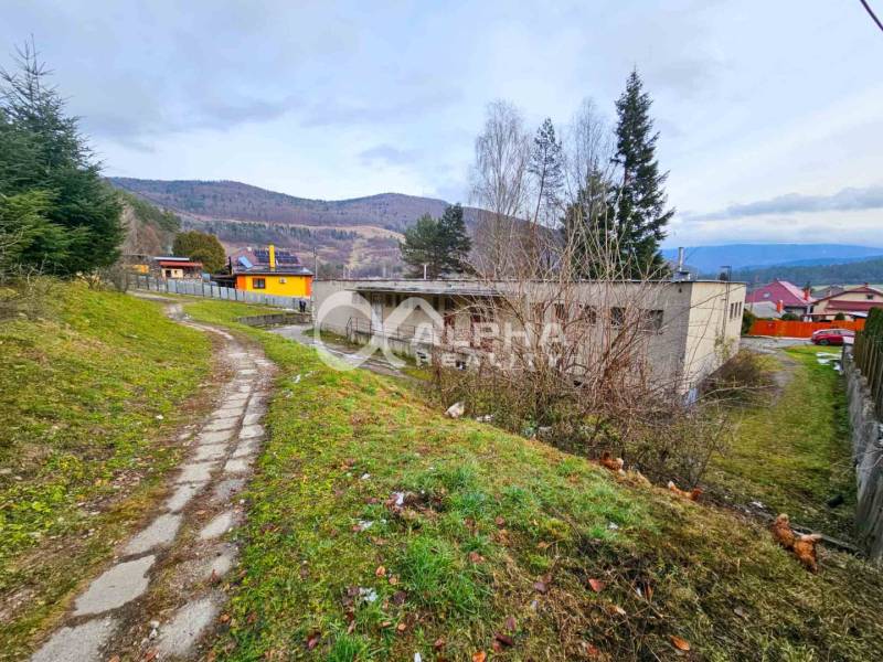 A building in Helcmanovce, surrounded by hills and a road with a lawn and trees.