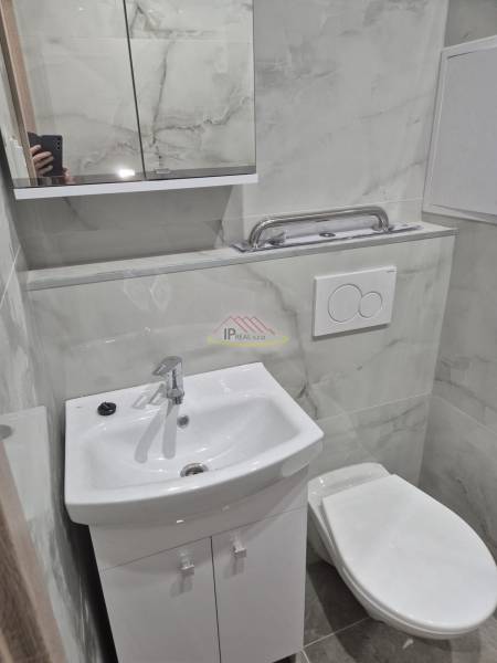 A bathroom in a studio apartment with a sink, mirror, and toilet, tiled with light tiles.