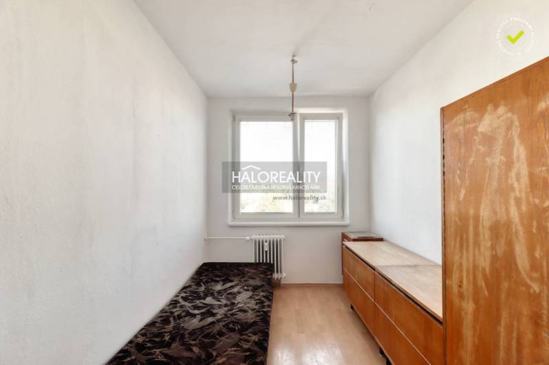 A bedroom in a two-room apartment with a window, a simple bed, and a wooden wardrobe.