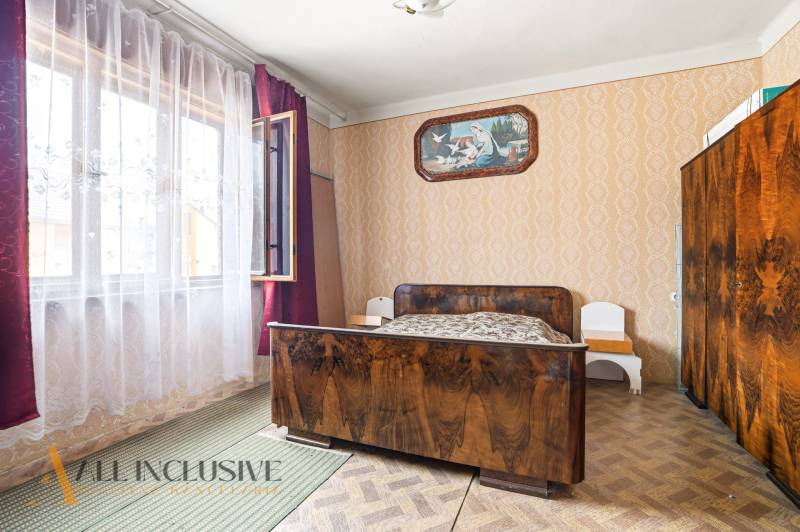 A room in a family house with a bed, a wooden-patterned floor, and patterned wallpaper.